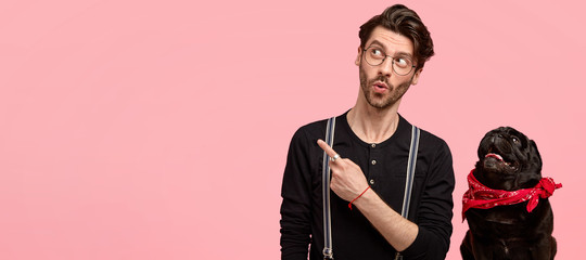 Shocked impressed guy has trendy haircut, bristle, wears round glasses, black shirt, stands near his favourite pet, indicates with index finger at copy space on right side, shows something incredible