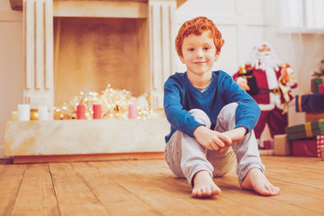 Cozy evening. Charming preteen boy sitting on the floor near the chimney in a beautifully decorated living room and waiting for the start of Christmas celebration