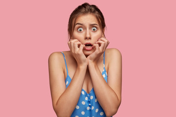 Wall Mural - Scared beautiful woman looks with great fear, keeps both hands near mouth, being frightened by something, dressed in polka dot dress, isolated over pink background. Emotional girl afraids of nightmare