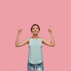 Wall Mural - Vertical shot of glad European woman smiles gladfully, points with both index fingers upwards, dressed in casual vest and leggings, shows where gym is situated isolated over pink background.