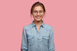 © wayhome.studio  - Studio shot of smiling glad woman has appealing appearance, smiles broadly, dressed in denim shirt, poses against pink background expresses positive emotions likes what she sees, rejoices new purchase