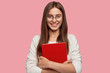 © wayhome.studio  - Waist up shot of pretty girl smiles pleasantly, carries red textbook closely, wears white casual jamper, models against pink background. Headshot of Caucasian lady student happy after having classes