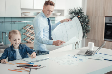 Wall Mural - Be creative. Occupied cute pleasant girl sitting in the kitchen near her father who overlooking the plan smiling and painting.