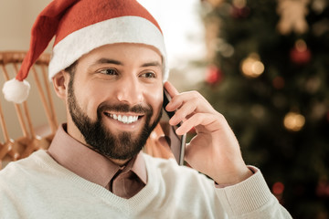 Wall Mural - Positive mood. Portrait of a cheerful delighted man smiling and talking on the phone while wearing a Santa hat