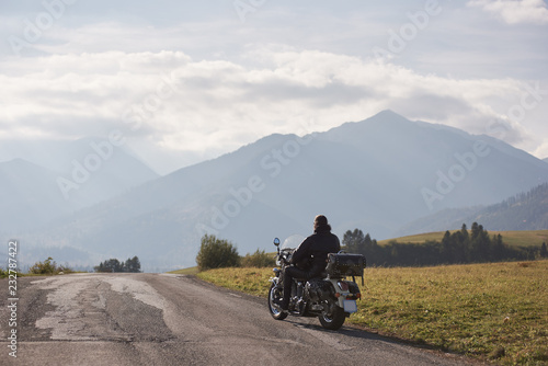 Obraz Dibond Tylny widok motocyklista w czarnej skórzanej kurtki krążownika jeździeckim rowerze wzdłuż drogi na zamazanej kopii przestrzeni tle piękny mgłowy pasmo górskie i błękitny chmurny niebo na jaskrawym pogodnym letnim dniu.