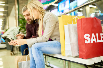 Side view portrait of adult couple sitting on bench in shopping mall and using smartphone, focus on red paper bag with word SALE