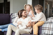 © fizkes - Young attractive married couple with little preschool children sitting on couch in living room at new modern wealthy house. Happy playful parents and joyful kids spending free time or weekend together