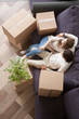 © fizkes - Young married couple embracing sitting together on couch in living room at their new house with unopened stuff in cardboard boxes, top above view. Buying of new real estate home or relocating concept