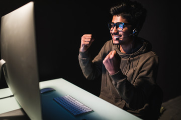 Ecstatic asian gamer boy rejoicing victory while playing video games on computer in dark room wearing headphones and using backlit keyboard