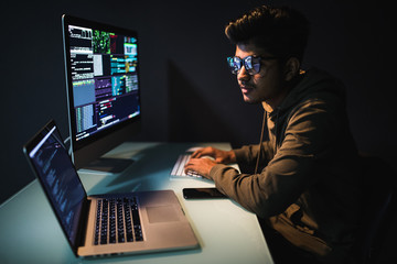 cybercrime, hacking and technology concept. indian male hacker in dark room writing code or using co