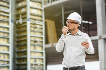 Male head engineer wearing white safety hardhat with walkie talkie and tablet inspecting construction site.