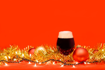Dark ale beer in a tulip glass with christmas baubles tinsel and lights on red background