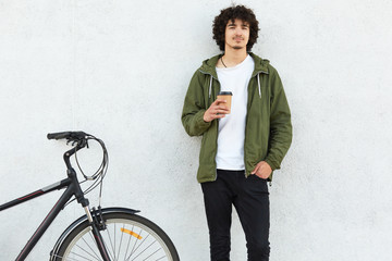 People, youth and lifestyle concept. Handsome young male dressed in fashionable outfit, drinks aromatic hot coffee, poses against white concrete wall near bicycle, ready to cover long distance