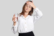 Tired overworked woman keeps hand on forehead, wears transparent glasses, black and white clothes, holds glasses of water, feels thirsty and fatigue, isolated over white background. Weariness