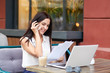 © sementsova321 - Cheerful brunette businesswoman has telephone conversation, satisfied with consultancy, talks with service operator, dressed in fashionable blouse, holds paper documents, works on laptop computer