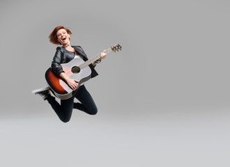 Wall Mural - Young woman musician with an acoustic guitar in hand jumping high on a gray background. He laughs and plays rock and roll loudly.