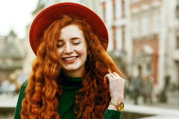 Close up portrait of young beautiful fashionable happy smiling redhead woman with freckles, very long curly hair, wearing green turtleneck, orange hat, golden wrist watch, posing in street. Copy space