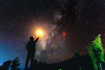 silhouette of a standing human looking milky way at the night time. light holds up over his head.