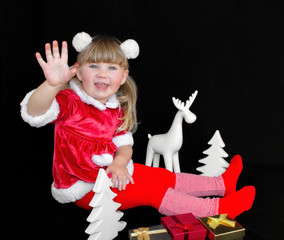 Little beautiful girl sitting on a black background in a Santa costume, with fur balls on her head, holding gifts in her hands and rejoices around her deer and Christmas trees. The concept of celebrat