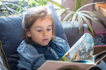 Poster - Little girl is reading a book in the living room on the couch