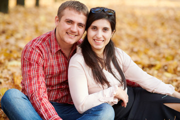 Couple is sitting on yellow fallen leaves in autumn city park. Bright yellow trees.