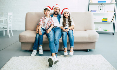 Wall Mural - Handsome father with him son and daughter. Happy young family in red santa hats spending time together at home while winter holidays