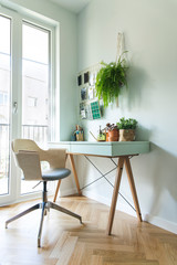 Modern scanidinavian interior of home office with design table, armchair, office accesories, big window and plants. Stylish and sunny space with wooden brown parquet.	