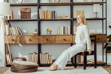 Wall Mural - adult blonde woman in glasses sitting near rack with books