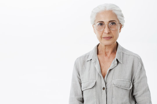 Studio shot of wise and intelligent skillful senior female doctotr in glasses with gray hair standing calm and confident against white background looking self-assured at camera