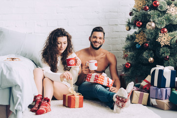 happy young couple with coffee cups sitting near gift boxes and christmas tree at home