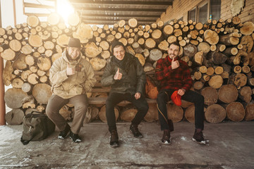 Best time. Finger up. Happy wokers lumberjacks wearing in jackets on the bench on the background of sawmill and warehouse of trees