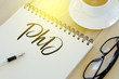 © MohamadFaizal - Top view of pen,sunglasses,a cup of coffee and notebook written with PhD on wooden background.