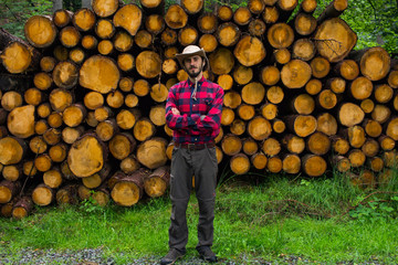 Portrait of lumberjack in forest, many big logs of pine on background. Young male hiker posing near the sawmeal in the pines forest.