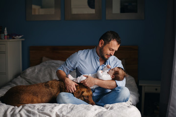 Portrait of middle age Caucasian father with newborn baby. Dog pet laying on bed. Man parent holding child in hands. Authentic lifestyle documenatry moment. Single dad family life.