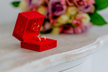 Two Golden Wedding Rings On A Wooden Table In A Red Wedding Box. With Some Flowers In The Background.