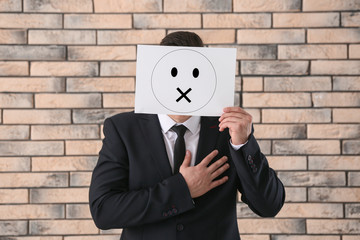 Businessman hiding face behind sheet of paper with drawn emoticon against brick wall