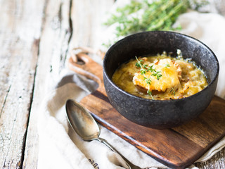 Traditional french onion soup with toast, cheese and fresh thyme in a black bowl on a wooden brown board on a rustic wooden background. Copy space