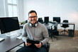 © bnenin - Portrait of a smiling young man with headset sitting in front of computer, looking at camera, in modern office.