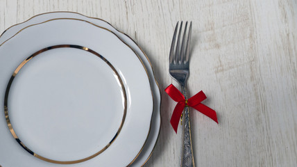 two white plates with a gilded cut edge and a fork with a red bow on a wooden background. festive table setting