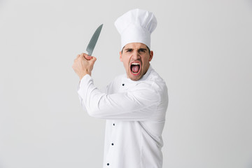 Wall Mural - Agressive screaming young man chef indoors isolated over white wall background holding knife.