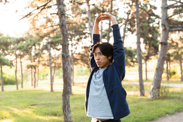 Wall Mural - Image of healthy asian guy in casual wear stretching his body, while doing sports in green park