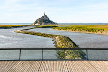 View Of The Mont Saint-Michel Tidal Island, Located In France On The Limit Between Normandy And Brittany, At High Tide With The Wooden Footbridge Over The Dam On The Couesnon River In The Foreground.