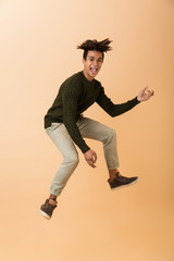 Wall Mural - Full length photo of joyous african american guy wearing sweater laughing and jumping, isolated over beige background