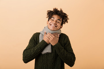 Wall Mural - Portrait of happy african american man wearing sweater and scarf touching throat, isolated over beige background