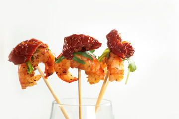 Grilled shrimps with the dried tomato and arugula on sticks, a canape stand in a glass on a white background