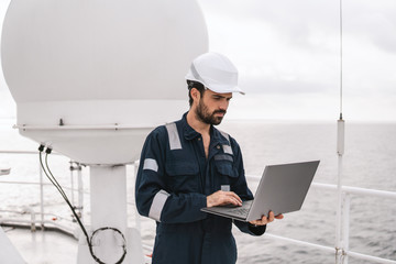 Marine service technician or serviceman near VSAT terminal on deck of vessel or ship. He is working on laptop or notebook