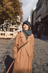 Wall Mural - Portrait of a young woman in brown coat and gray cap. Woman posing outdoors on the street at bright sunny autumn day