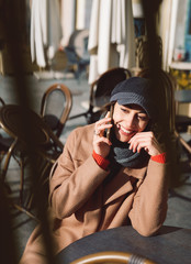 Wall Mural - Portrait of a young woman in brown coat and gray cap using phone. Smiling cheerful woman is sitting outdoors at the table in the street cafe at bright sunny autumn day