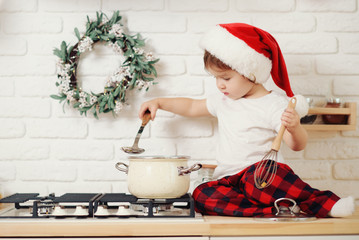 Cute little girl in santa hat, preparing cookies in the kitchen at home. Sits on the kitchen table and helps mom prepare a festive Christmas dinner