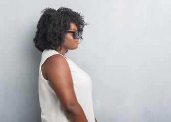 Poster - Young african american plus size woman over grey grunge wall wearing fashion sunglasses looking to side, relax profile pose with natural face with confident smile.